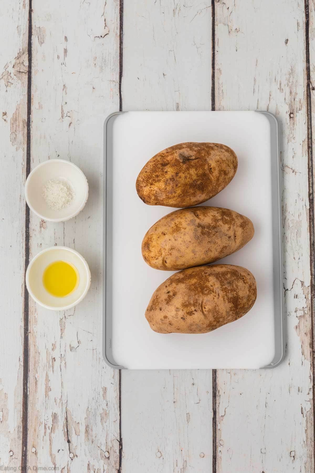 A white cutting board holds three whole russet potatoes placed in a row. On the left side of the cutting board are two small white bowls, one containing salt and the other olive oil—perfect ingredients for preparing air fryer baked potatoes—on a rustic white wooden surface.