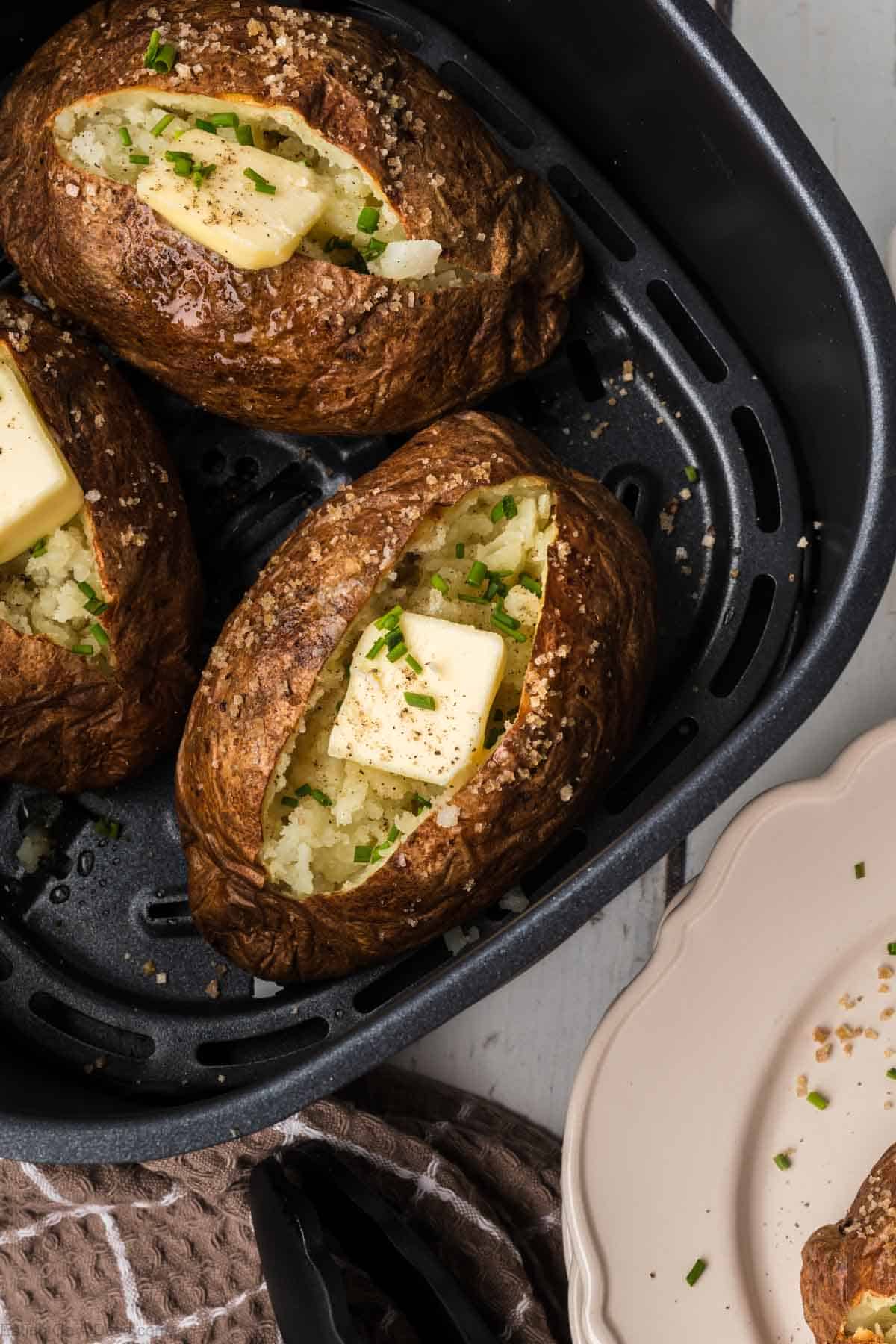 Three air fryer baked potatoes are shown, each sliced open with a pat of butter melting inside. Chopped chives are sprinkled as a garnish. A beige plate is partially visible in the background, while a brown towel rests near the air fryer basket.