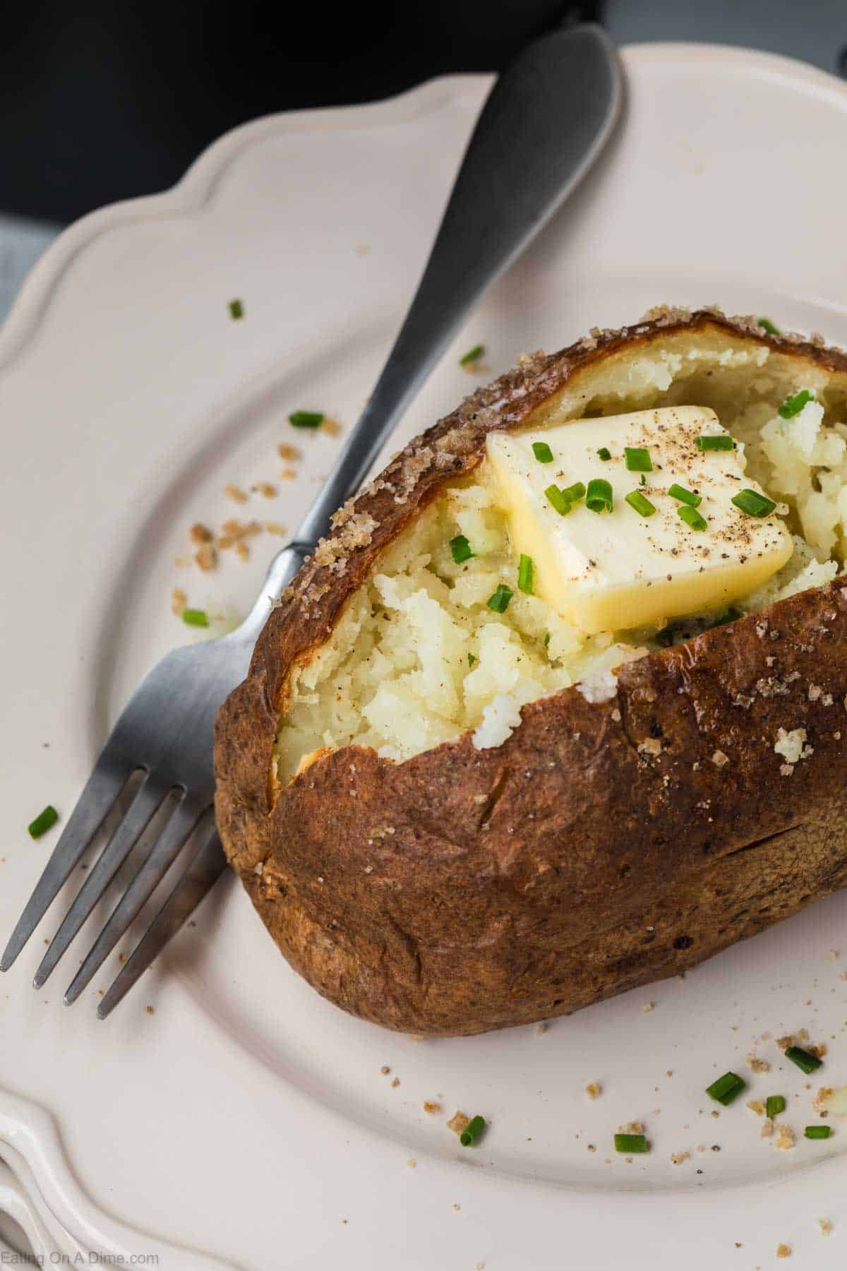 An air fryer baked potato split open, topped with a slice of melting butter, freshly ground black pepper, and chopped chives. It is served on a white plate with a fork resting beside it.