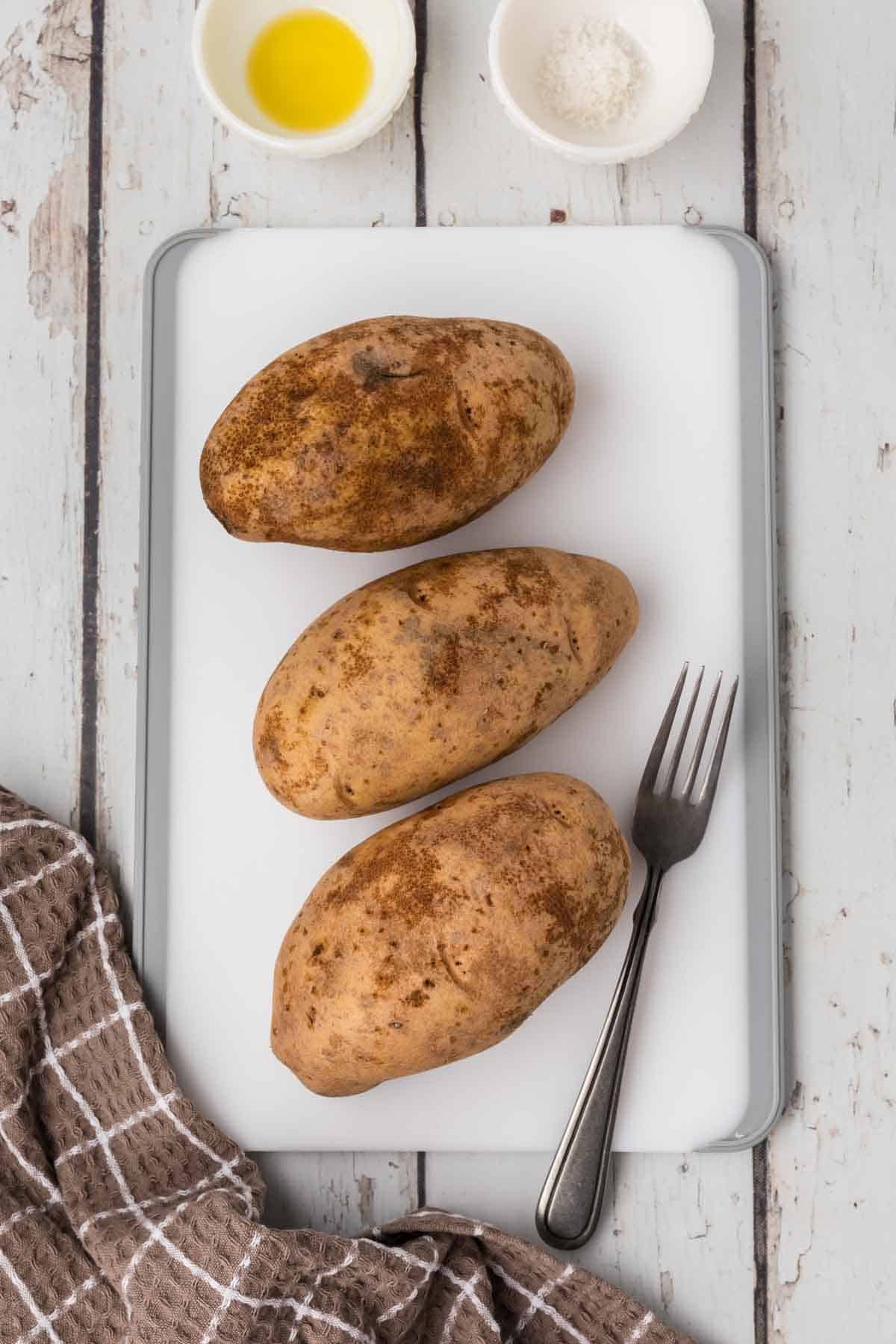 Three unpeeled potatoes are placed on a white cutting board with a fork beside them, ready for their turn as air fryer baked potatoes. Next to the cutting board is a small cup of oil, a small bowl of salt, and a brown checkered cloth. The scene is set on a rustic white wooden surface.