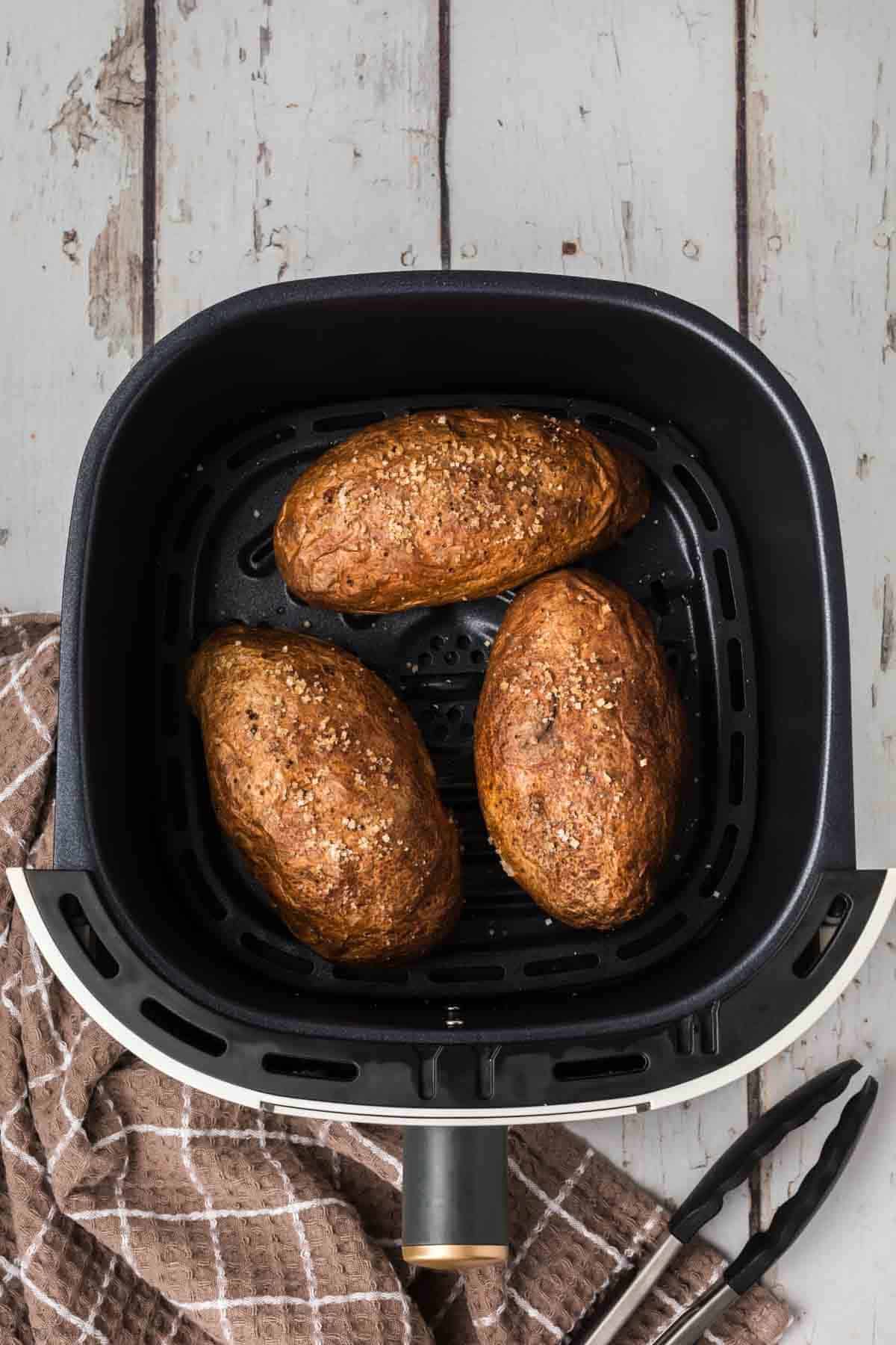 An air fryer basket containing three perfectly baked potatoes seasoned with coarse salt and herbs. The basket is placed on a rustic wooden surface next to a pair of black tongs and a brown and white checkered kitchen towel, showcasing the ultimate air fryer baked potato experience.
