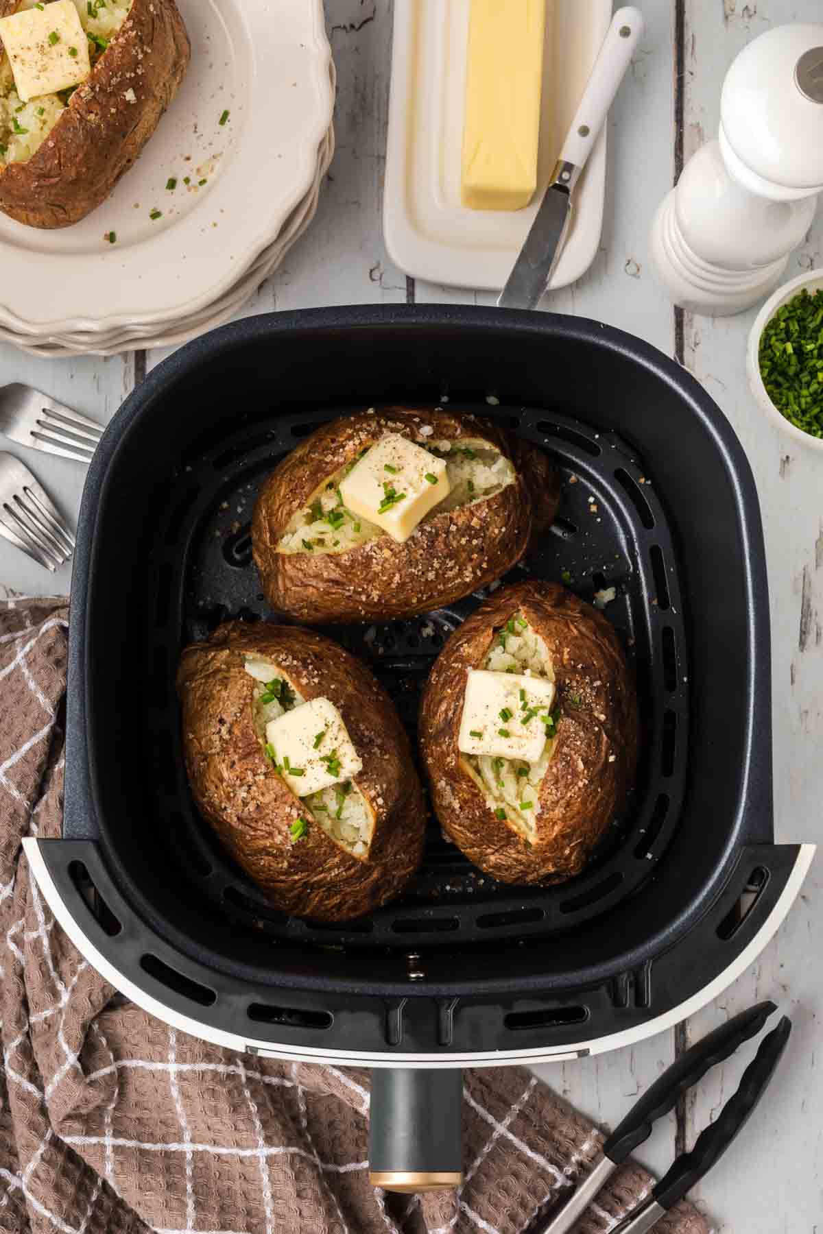 Three air fryer baked potatoes topped with butter and chopped herbs are in an air fryer. Nearby are forks, a knife, a brown kitchen towel, white plates with another potato, a stick of butter on a dish, a pepper grinder, and a bowl of chopped herbs.