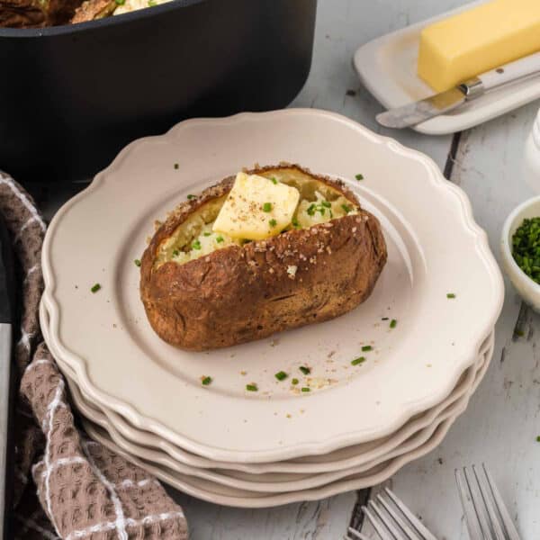 A crispy air fryer baked potato topped with a pat of butter, chives, and seasoning sits on a stack of white plates. A block of butter, a knife, and a napkin are in the background, along with a small bowl of chopped chives and forks.