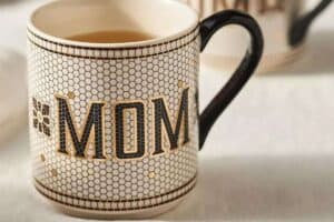 A ceramic mug with a honeycomb pattern showcases the word "MOM" in bold, black and gold letters. Perfect for Mother's Day gifts, it's filled with a hot beverage, and its handle is black. The softly blurred background adds a touch of elegance.