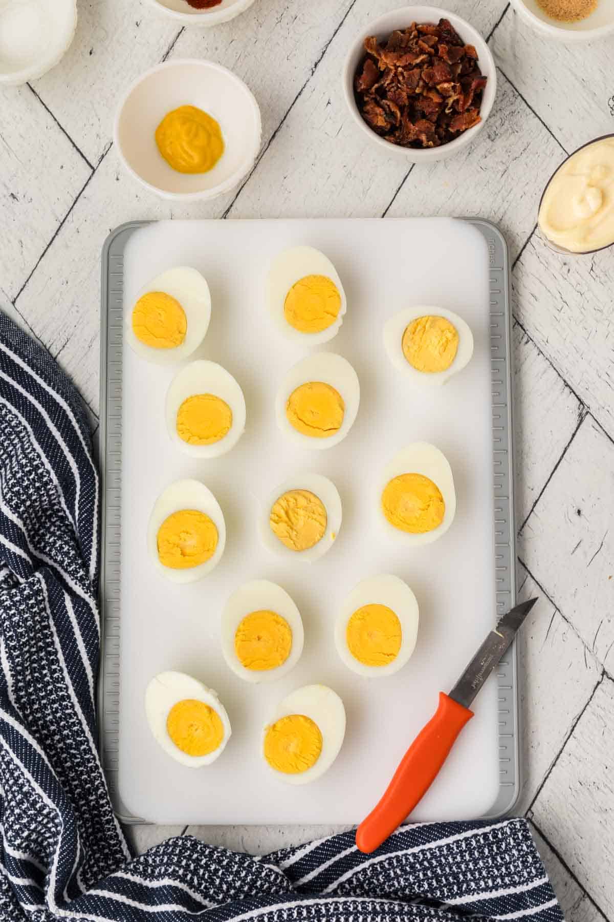 Hard boiled eggs cut in half on a cutting board with bowls of crushed bacon, mustard, and mayo on the side