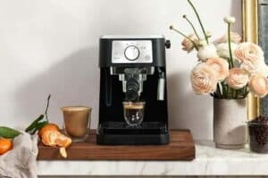 A black espresso machine on a wooden tray with a glass cup of espresso, perfect for thoughtful Mother's Day gifts. Nearby are a cup of coffee, peeled clementine, and pink flowers in a vase on a marble surface against a white wall.