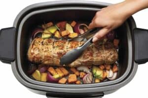 A person uses tongs to hold a cooked roast in a slow cooker, perfect for Mother's Day gifts. The roast is surrounded by diced vegetables like sweet potatoes, onions, and zucchini, with sprigs of rosemary adding aromatic flair.