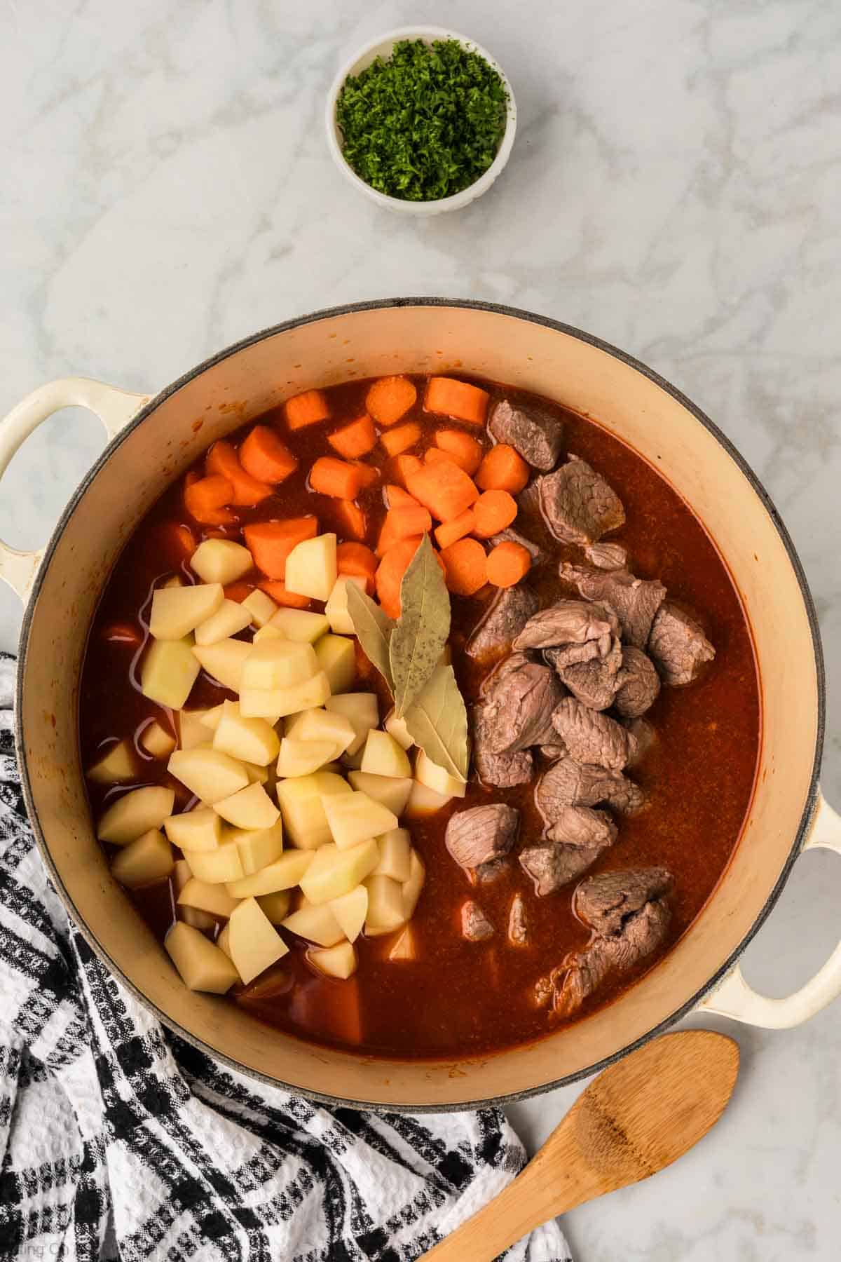 Diced potatoes and carrots and beef stew in a large pot topped with bay leaves in broth in a large pot