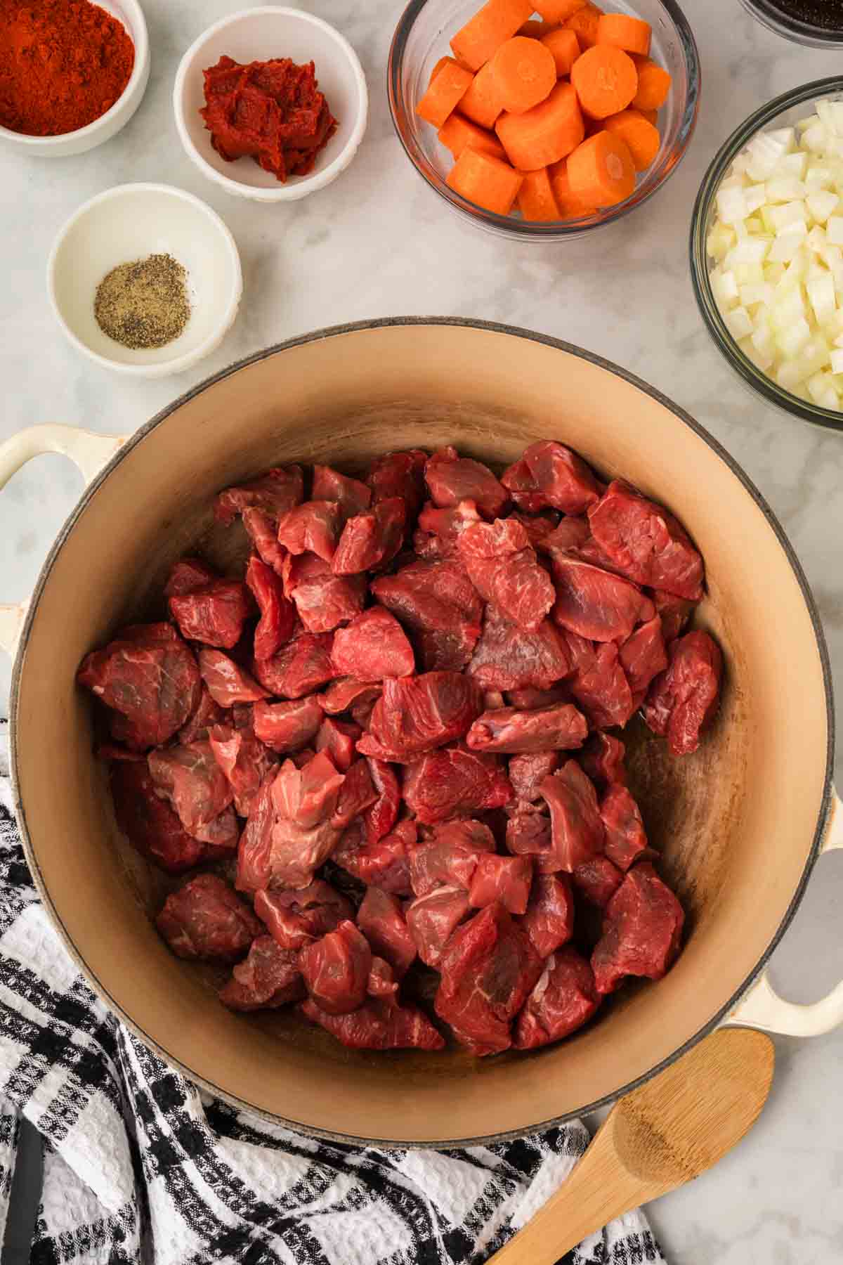 Beef Stew placed in a large pot, with a diced carrots, onions, paprika, tomato paste and seasoning in bowl on the side