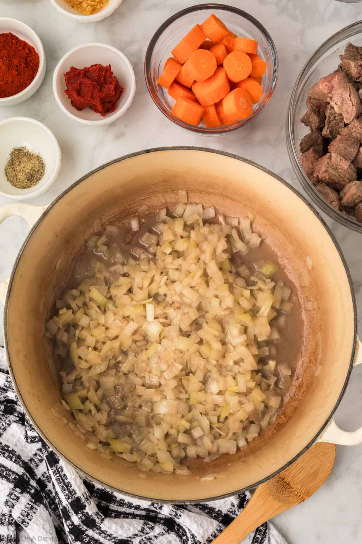 Cooked diced onions in a large pot with a side of diced carrots, cooked beef stew, tomato pasta, seasoning and paprika