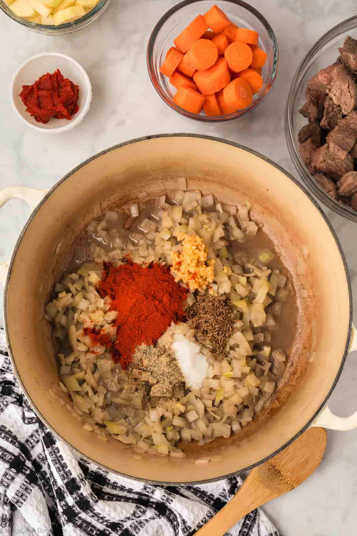 Diced onions in a large pot with paprika, minced garlic and seasoning in a large pot with a side of diced carrots, cooked beef stew 