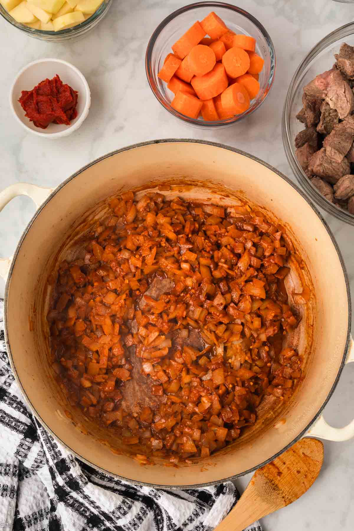 Tomato paste mixed with diced onions and seasoning in a large pot with a small bowl of carrots and cooked beef stew