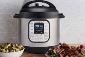 A stainless steel multi-function pressure cooker sits on a kitchen counter, its digital timer reading 00:18. Beside it, a plate holds grilled meat and roasted Brussels sprouts. Perfect for preparing meals with ease, this appliance makes an ideal Mother's Day gift against the backdrop of a white geometric tiled wall.