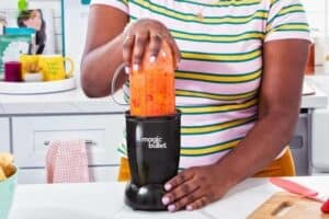 A person wearing a striped shirt uses a Magic Bullet blender to mix an orange smoothie in the kitchen, perhaps perfecting a recipe for Mother's Day gifts. Various kitchen items and a "Good Vibes Only" sign add charm to the setting.