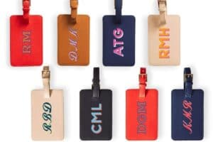 A variety of colorful luggage tags, perfect for Mother's Day gifts, each featuring initials like RM, DMK, and ATG. These tags come in leather finishes with contrasting embroidered letters and include a strap and buckle for easy attachment.