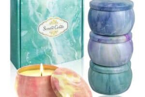 A set of scented candles in colorful, marbled tins makes perfect Mother's Day gifts. One candle is lit with a warm flame, while three other unopened candles are stacked beside it. The background features a decorative box labeled "Scented Candle" with a blue and green marbled design.