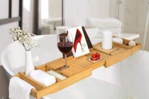 A bathtub caddy tray holds a glass of red wine, a book, a lit candle, and a small bowl of strawberries, crafting the ultimate Mother's Day gift. The tray spans across the filled bathtub for ultimate relaxation. A vase with white flowers adorns the tub's edge alongside a rolled towel.