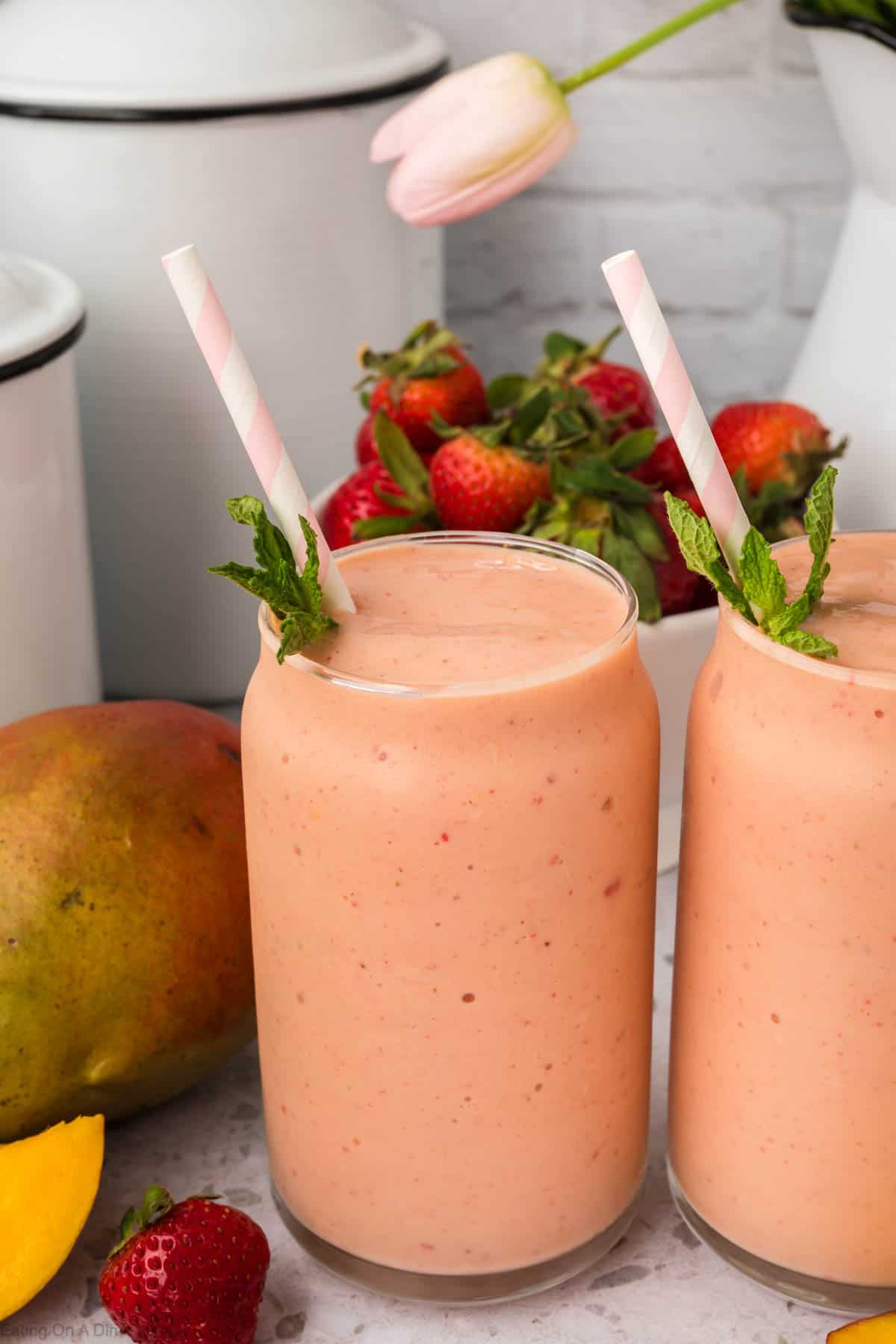 Strawberry Mango Smoothie in two glasses topped with fresh mint and a straw with a side of fresh strawberries and mango 