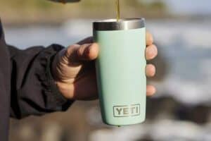 A person in a black jacket holds a light green Yeti tumbler, perfect for Mother's Day gifts, as liquid is being poured into it. The background is blurred, showcasing an inviting outdoor setting.