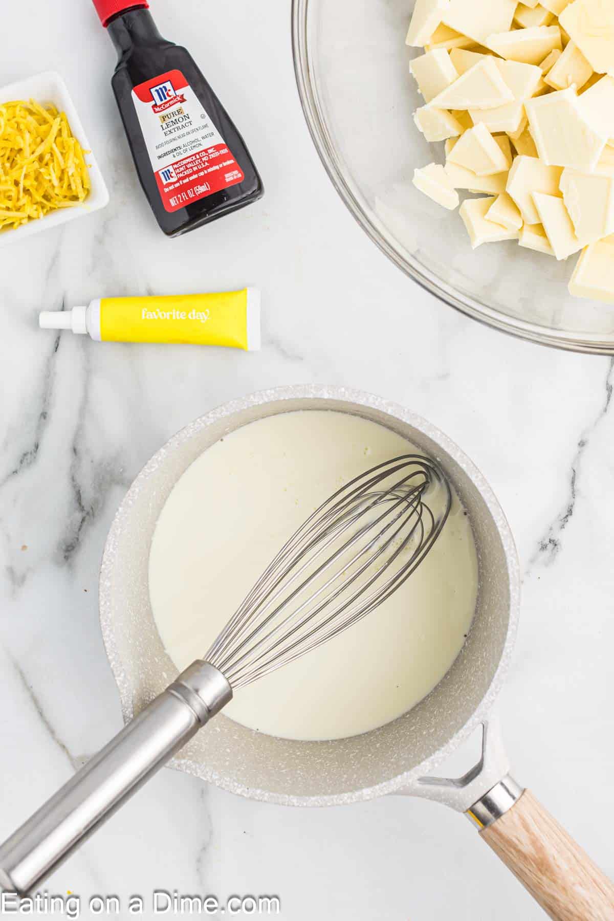 Heavy Cream with a whisk in a saucepan and a bowl of white chocolate pieces with lemon zest strips in a bowl, yellow food coloring and lemon extract on the side