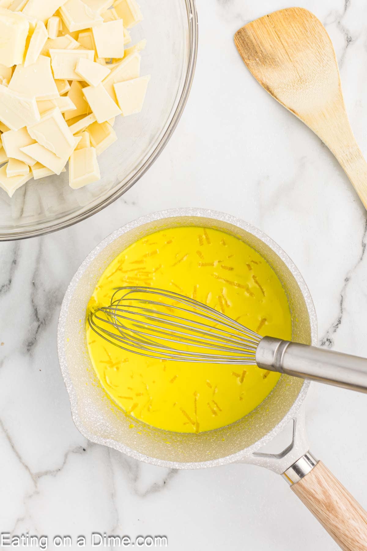 Lemon extract and lemon zest mixed with the heavy cream in a saucepan with a bowl of white chocolate pieces on the side