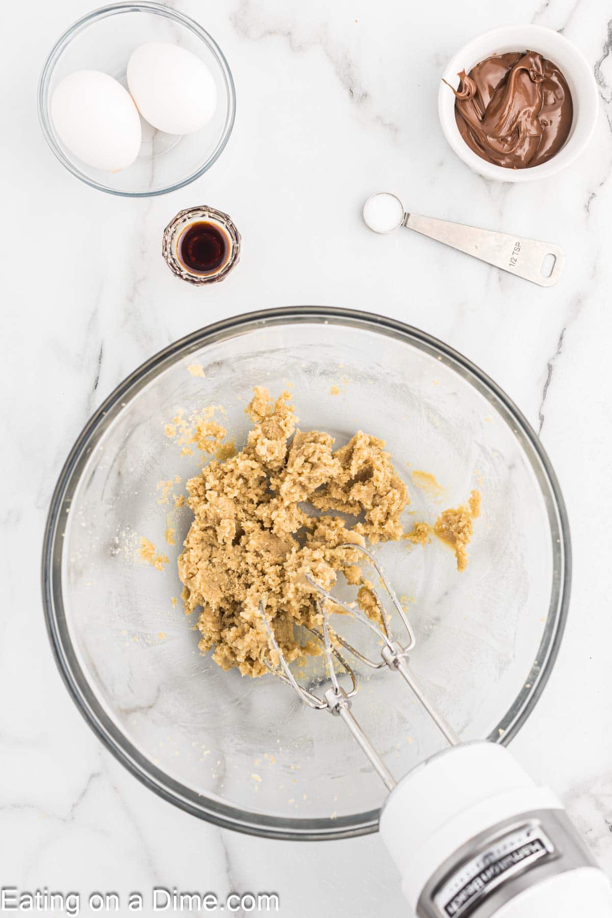 Beating butter and brown sugar in a bowl with a hand mixer with a small bowl of eggs, vanilla and Nutella on the side