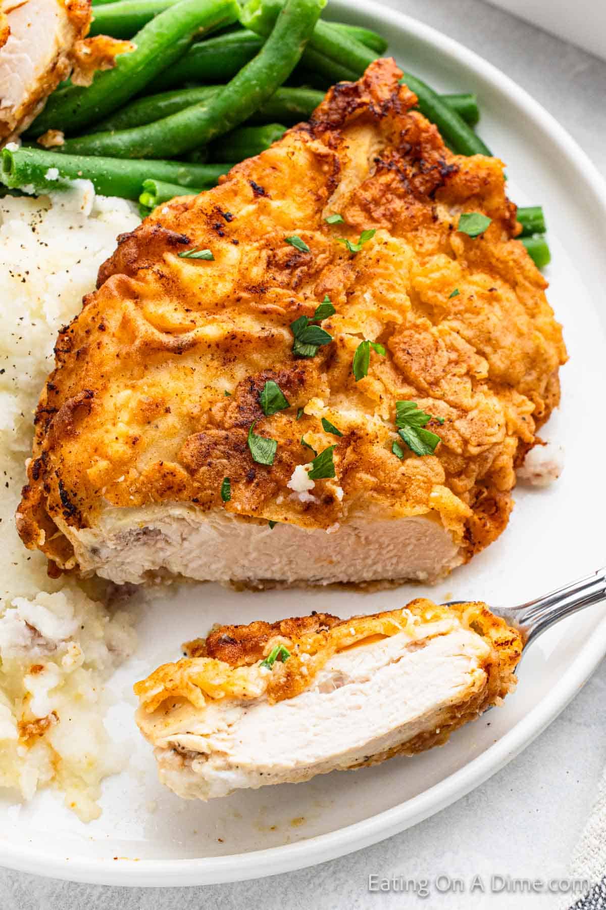 Fried chicken breast with a bite off the end that is on the fork with a side of mashed potatoes and green beans