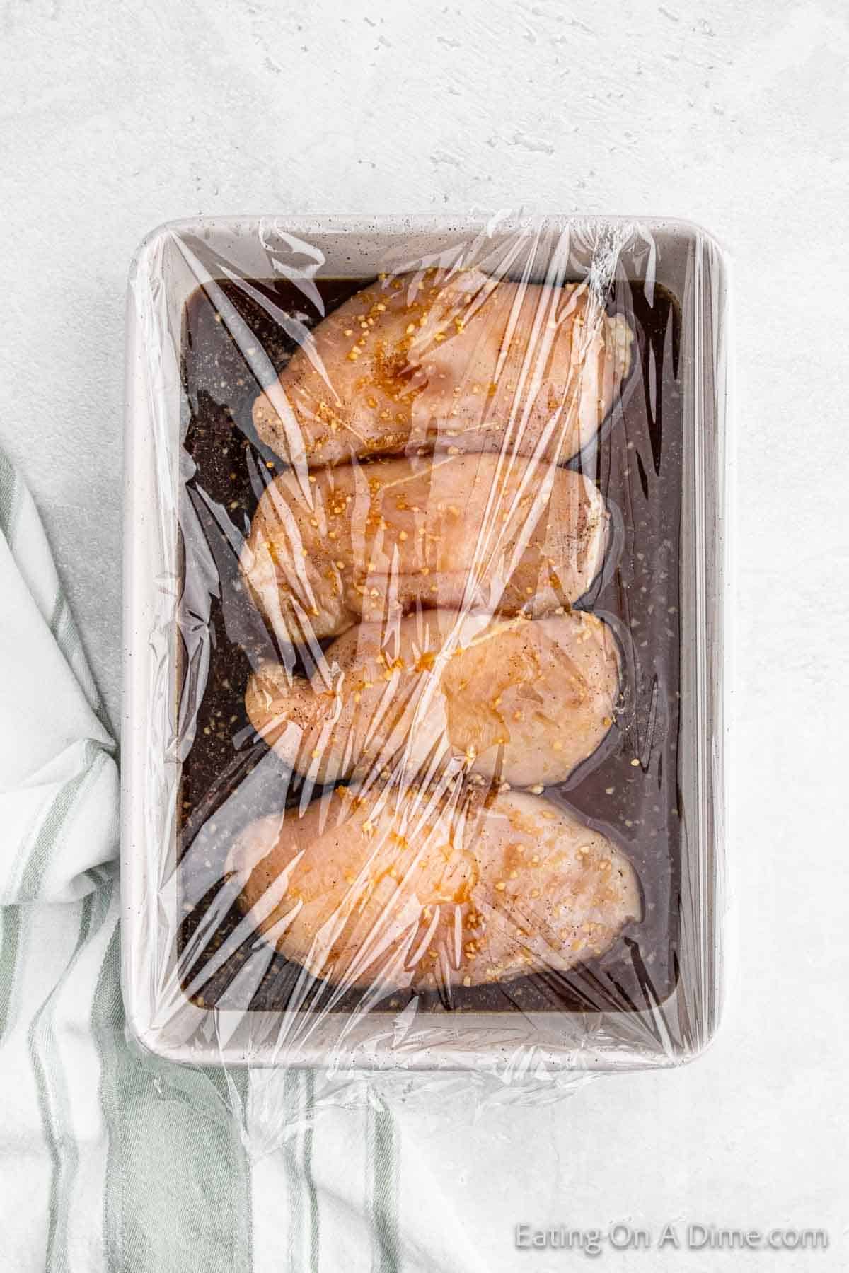 Placing chicken breast in a baking dish with the marinade covered with a plastic wrap