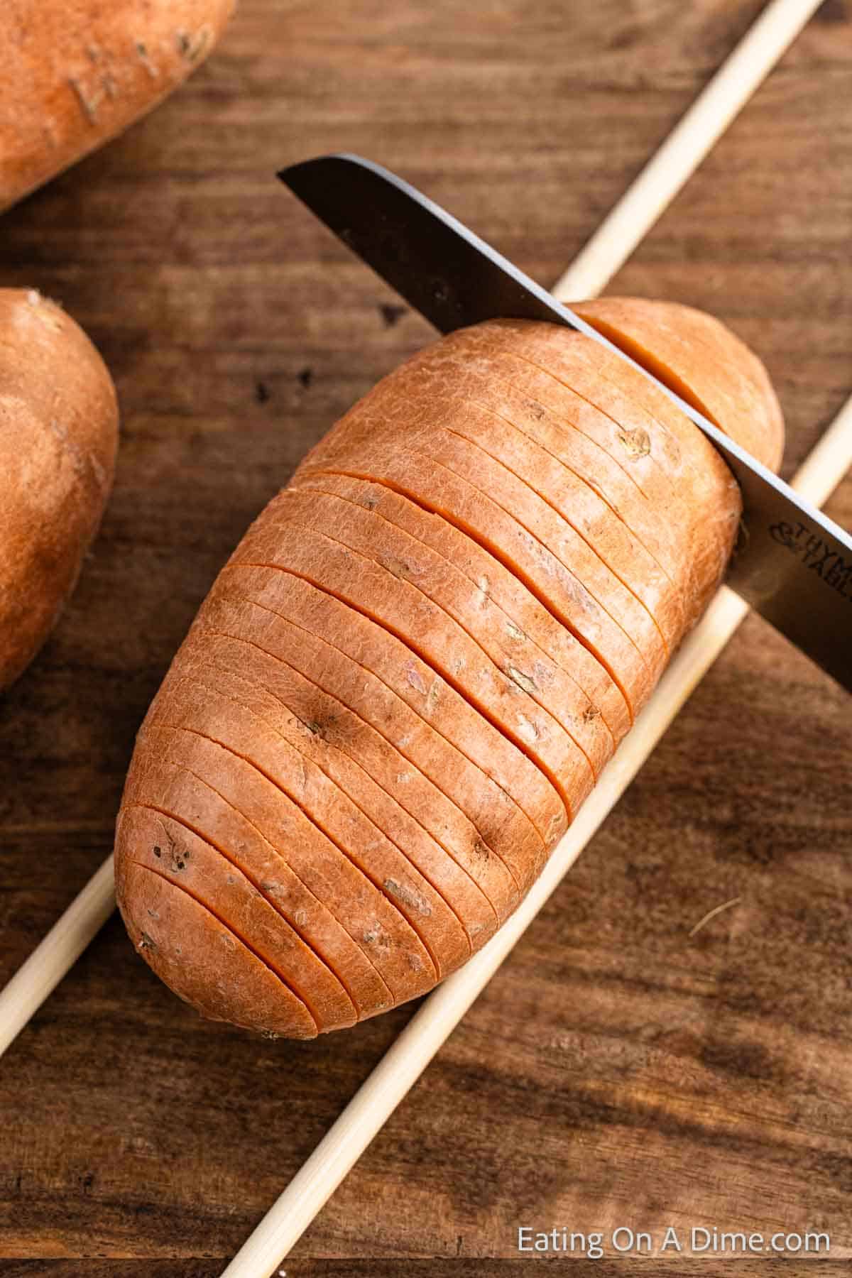 Slicing a whole sweet potato with a knife into small slits with wooden skewers on the side on a cutting board