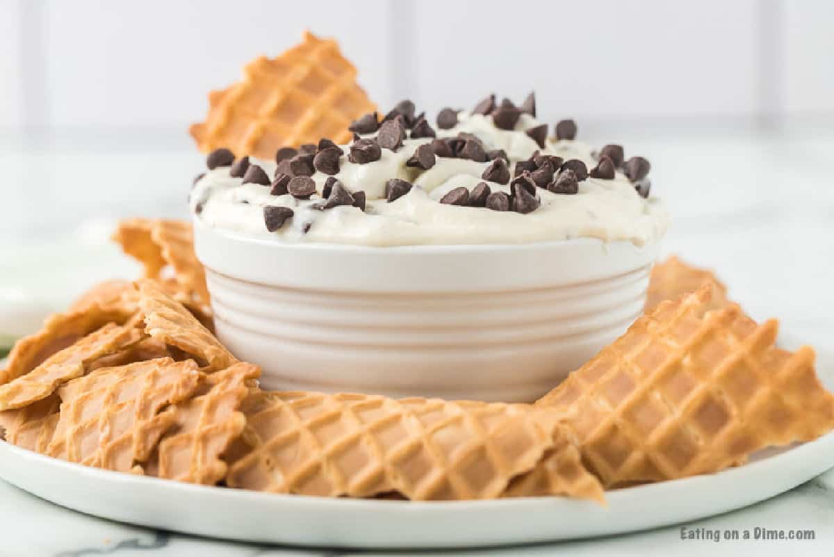 Cannoli Dip in a bowl topped with chocolate chips on a serving platter with pieces of cones on the side