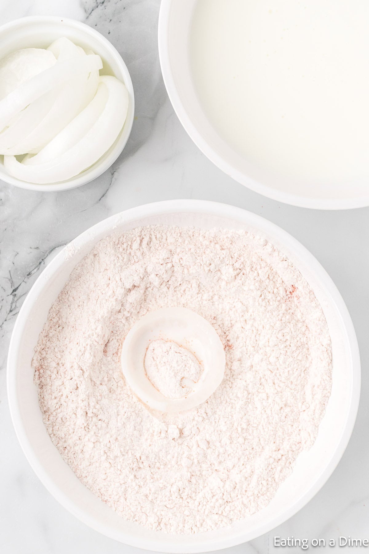 Bowl of onion slices, bowl of flour with onion on the flour, and a bowl of milk