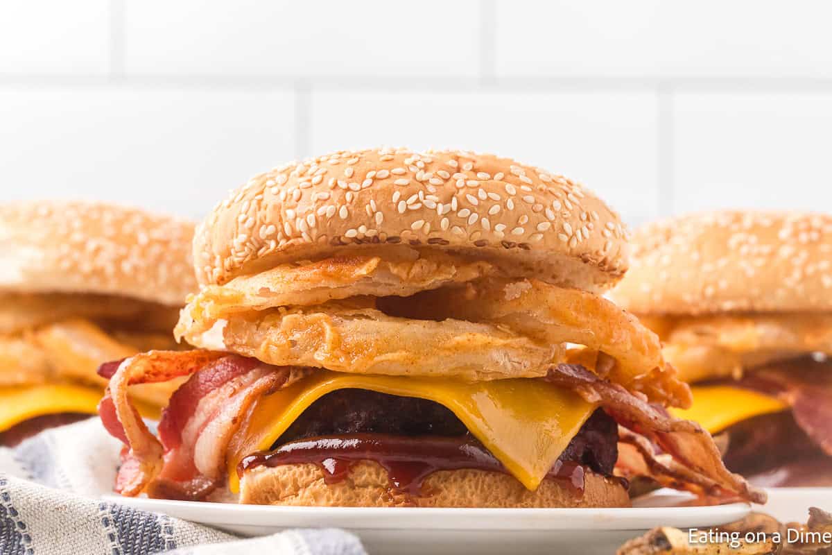 Hamburger that has slice tomato, green lettuce, crispy onion rings, bacon slices, slice of cheese and hamburger beef patties and BBQ Sauce all between seeded hamburger buns with the same burger in the background