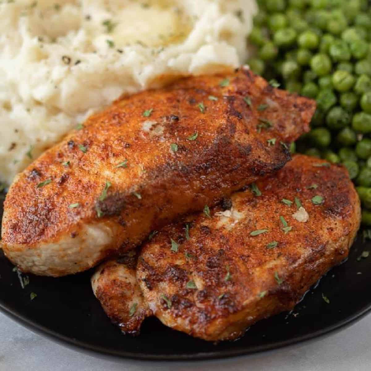 Close up image of pork chops on a black plate with a side of mashed potatoes and green peas. 