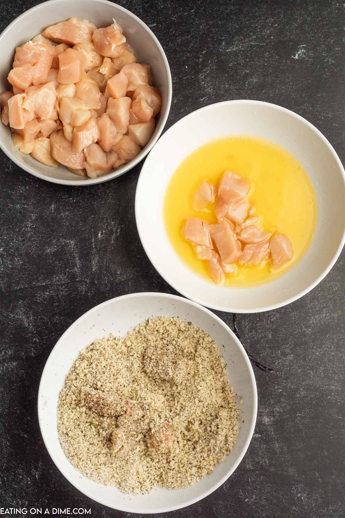 Bowl of bite size chicken, a bowl of melted butter with bite size chicken in it and a bowl of a bread crumb mixture with chicken in it