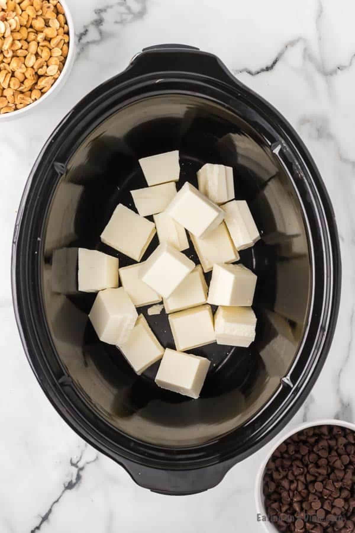 Almond bark pieces in the slow cooker with bowls of peanuts and chocolate chips