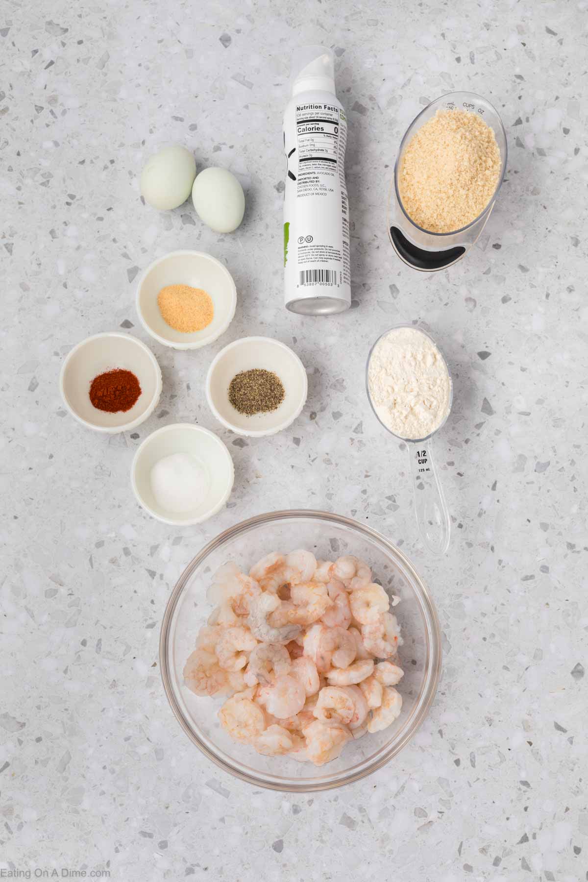 Ingredients for a popcorn shrimp recipe arranged on a countertop, including a bowl of raw shrimp, two eggs, bowls of seasoning, flour, breadcrumbs, and a can of cooking spray. The seasonings appear to include salt, black pepper, paprika, and garlic powder. Perfect for the air fryer.