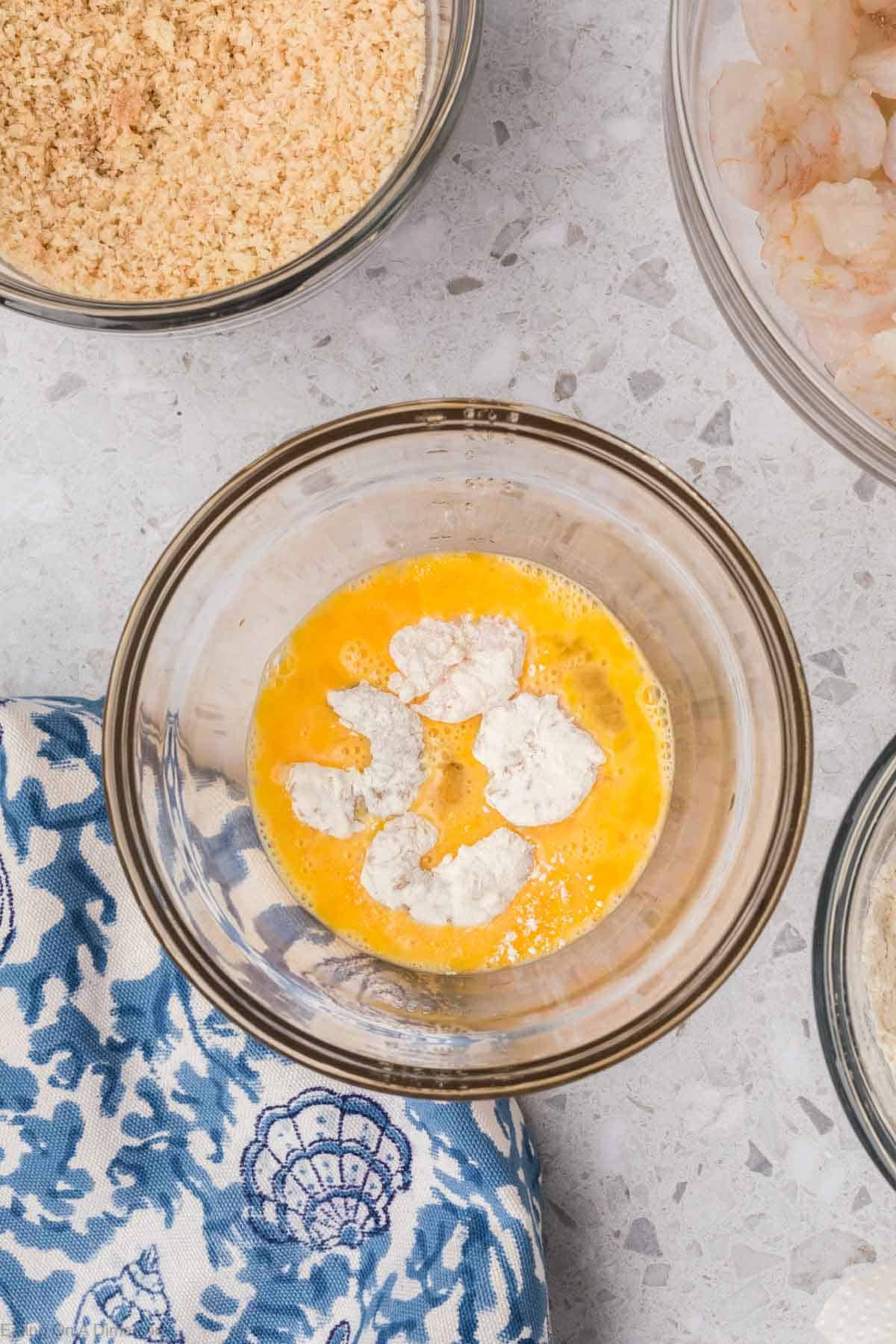 A bowl of raw shrimp is dipped in beaten eggs. Surrounding the bowl are other ingredients for breaded popcorn shrimp, including breadcrumbs and flour in additional bowls. A blue and white patterned cloth lies beside the bowls on a light textured surface. Perfect for the air fryer!