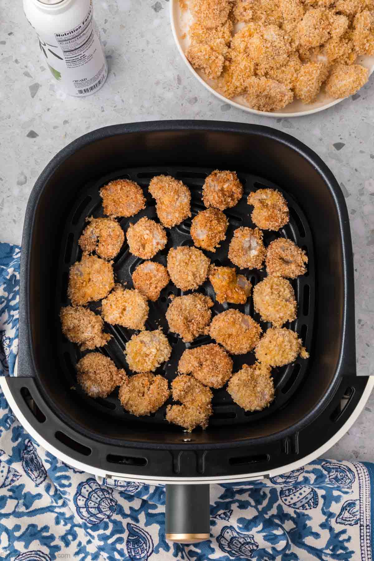 Breaded cauliflower pieces are spread inside an air fryer basket, ready for cooking. A plate with more breaded cauliflower is placed next to the air fryer, reminiscent of preparing Air Fryer Popcorn Shrimp. The background features a blue patterned cloth and a countertop.