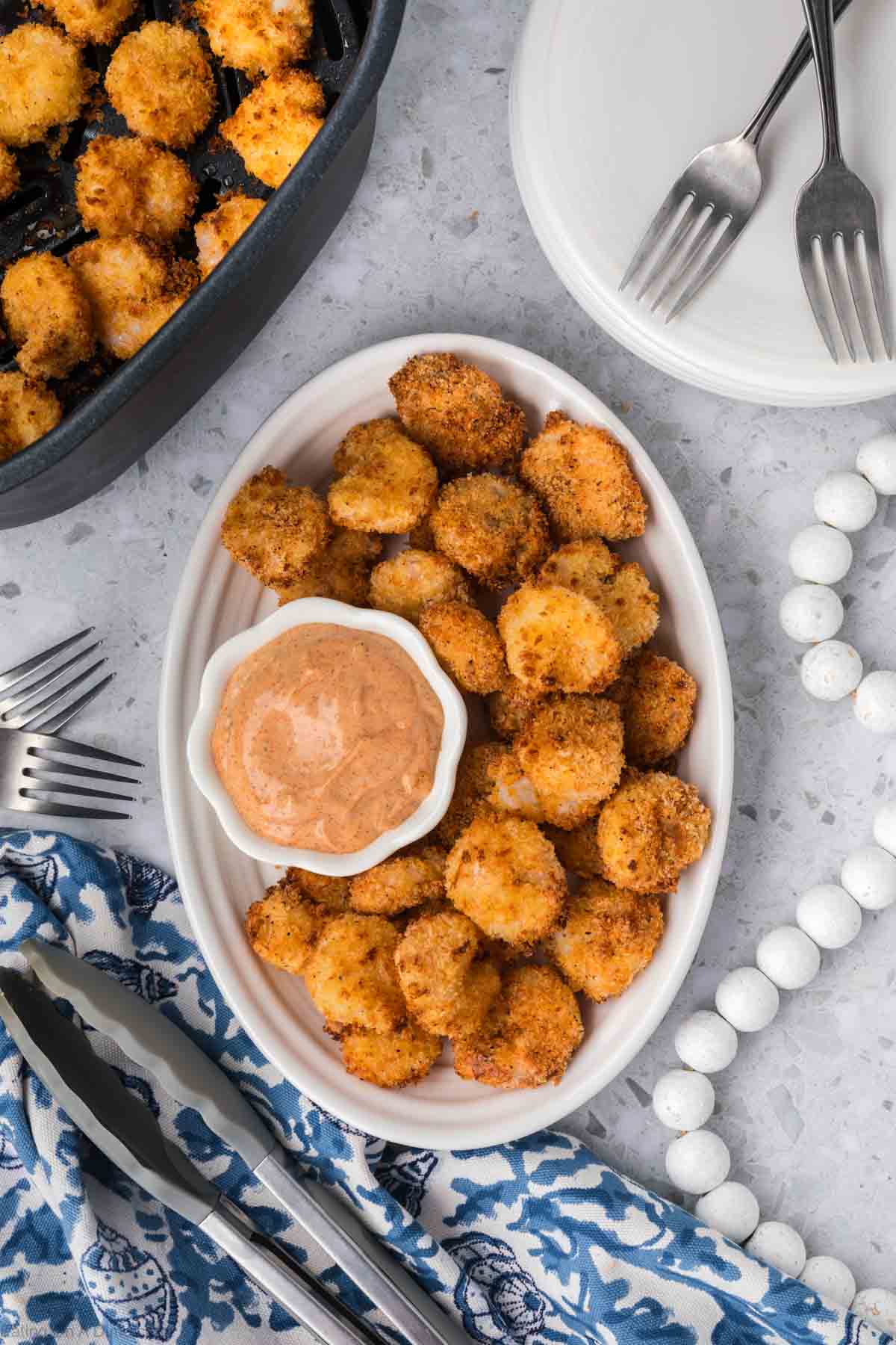         A white oval platter filled with crispy, golden-brown breaded bites of popcorn shrimp alongside a small white bowl of dipping sauce. The platter is on a light-colored surface with a blue printed napkin, forks, and tongs nearby. A dark dish in the top left corner holds more breaded bites.