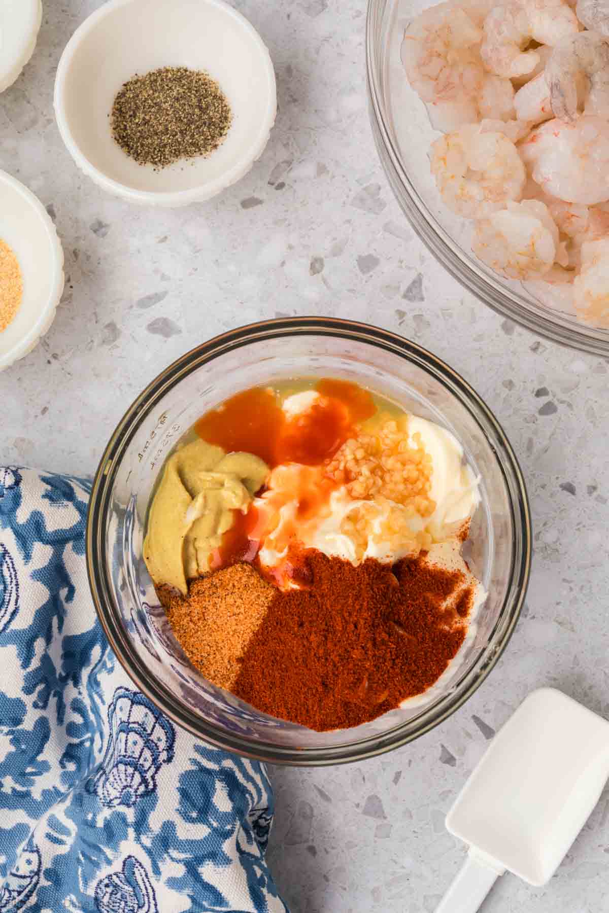 A bowl containing various ingredients, including mustard, paprika, mayonnaise, hot sauce, and minced garlic, is on a kitchen countertop. Nearby are a bowl of peeled shrimp ready to be transformed into crispy popcorn shrimp using an air fryer, a blue patterned kitchen towel, and small bowls of black pepper and garlic powder.