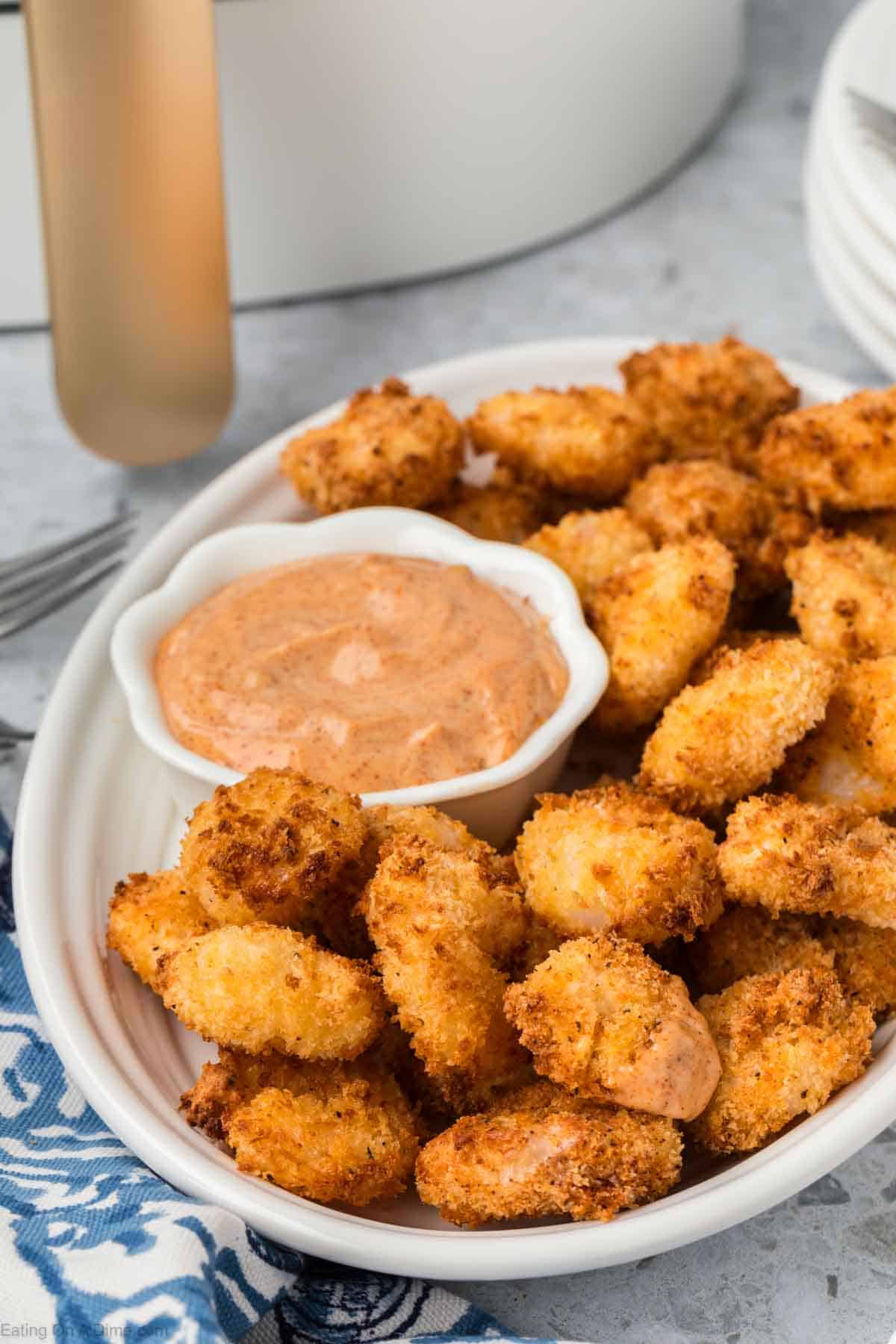 A white plate filled with crispy, golden-brown, air fryer popcorn shrimp is placed on a gray surface. In the center of the plate is a small white bowl containing a creamy, orange dipping sauce. A blue and white cloth is partially visible on the left side of the plate.