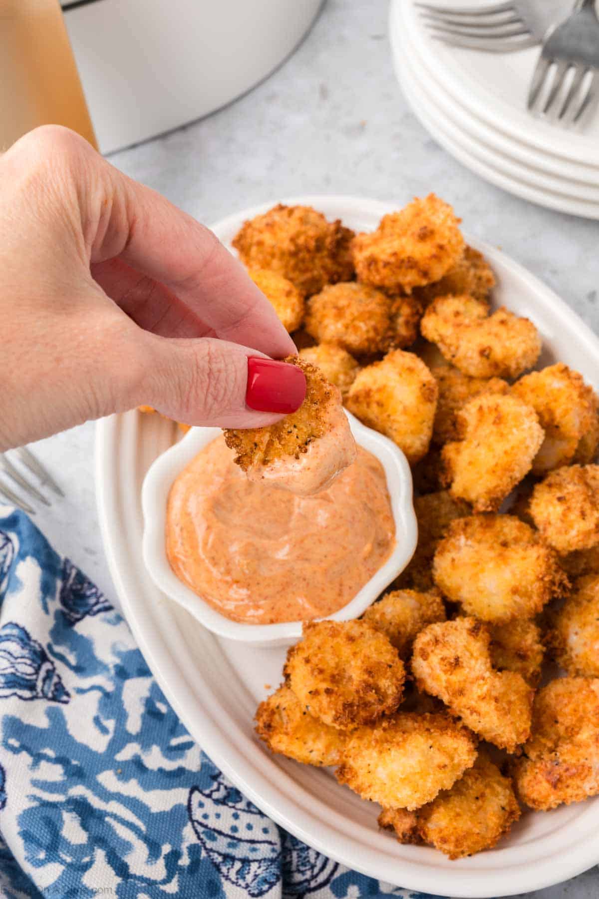 A hand with red nail polish is dipping a piece of air fryer popcorn shrimp into a small bowl of creamy orange dipping sauce. The shrimp is on a white oval platter, which is placed on a marble surface alongside a blue and white patterned cloth.