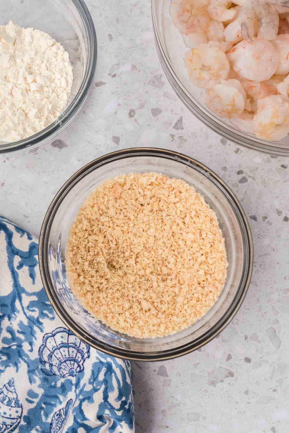 Top-down view of ingredients on a countertop: a bowl of bread crumbs, a bowl of flour, and a bowl of shrimp, perfect for making popcorn shrimp. A blue and white patterned cloth is placed nearby. Explore air fryer recipes for the best results.