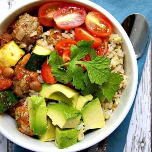 A vibrant bowl of food containing brown rice, avocado chunks, grape tomato halves, meatballs, zucchini pieces, and garnished with fresh cilantro. This delightful example from Rice Bowl Recipes is placed on a wooden surface alongside a blue napkin and a spoon.