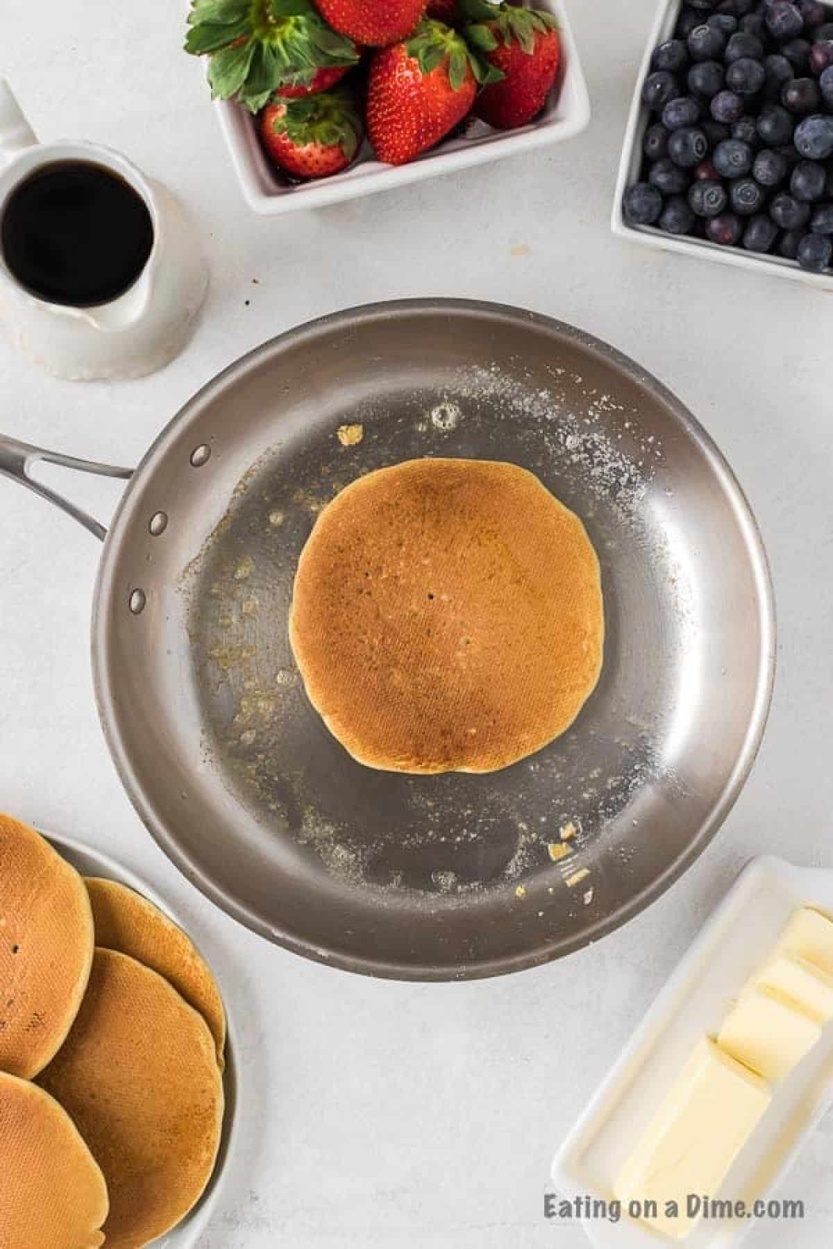 Cooked pancake in a large skillet with a side of a plate with pancakes, slice butter and blueberries