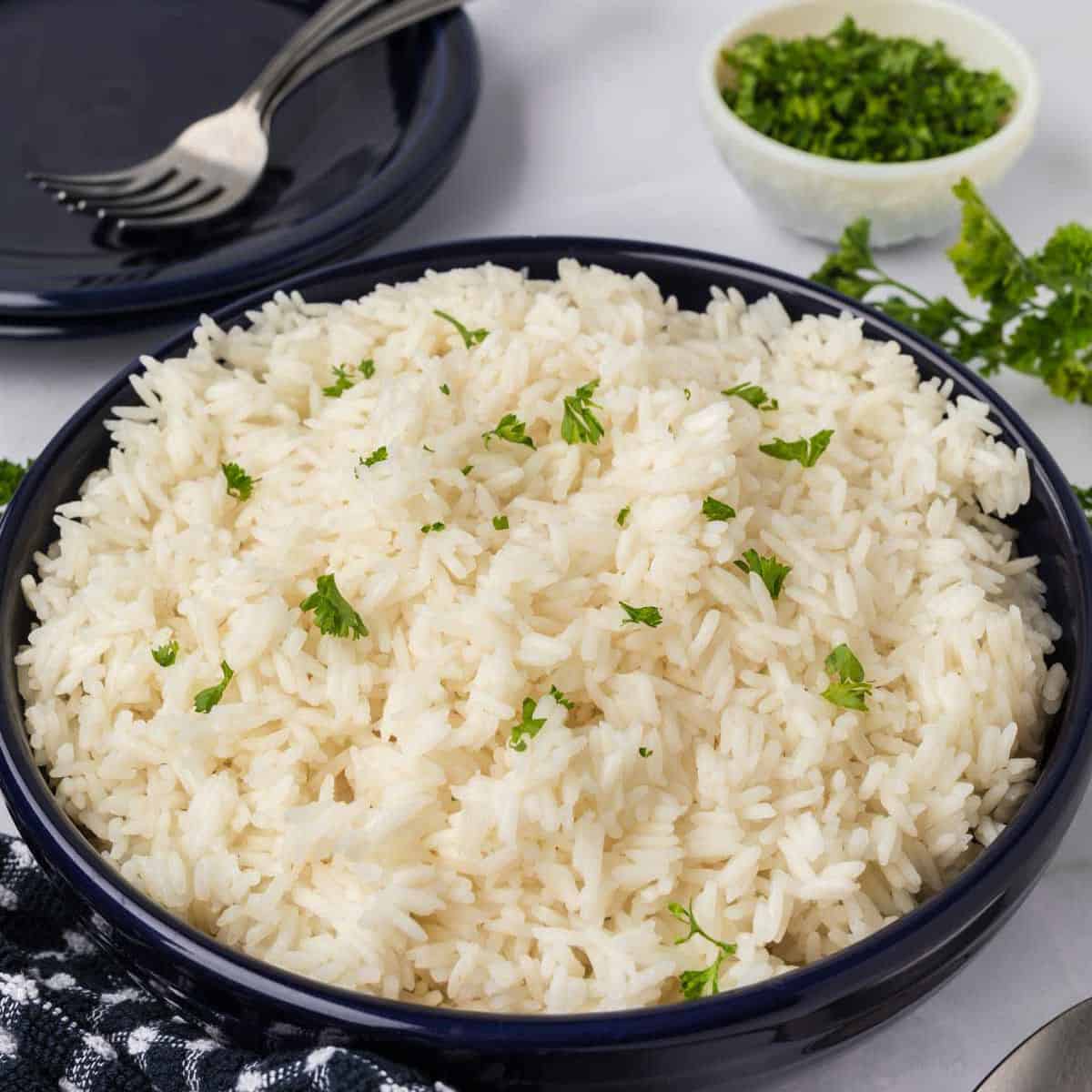 A bowl of fluffy Jasmine rice garnished with chopped herbs is placed on a table. A checkered cloth is partially visible in the foreground. There is also a small bowl of additional chopped herbs and an empty dark plate with a fork and spoon in the background.