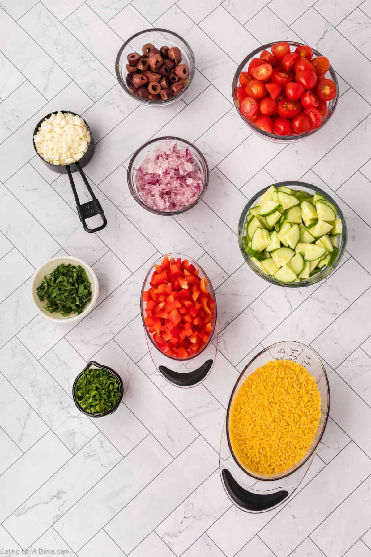 Overhead view of various chopped ingredients in bowls on a white tile surface. Includes cherry tomatoes, cucumber, red onion, black olives, feta cheese, red bell pepper, chopped cilantro, chopped jalapeños, cooked yellow rice, and orzo pasta.