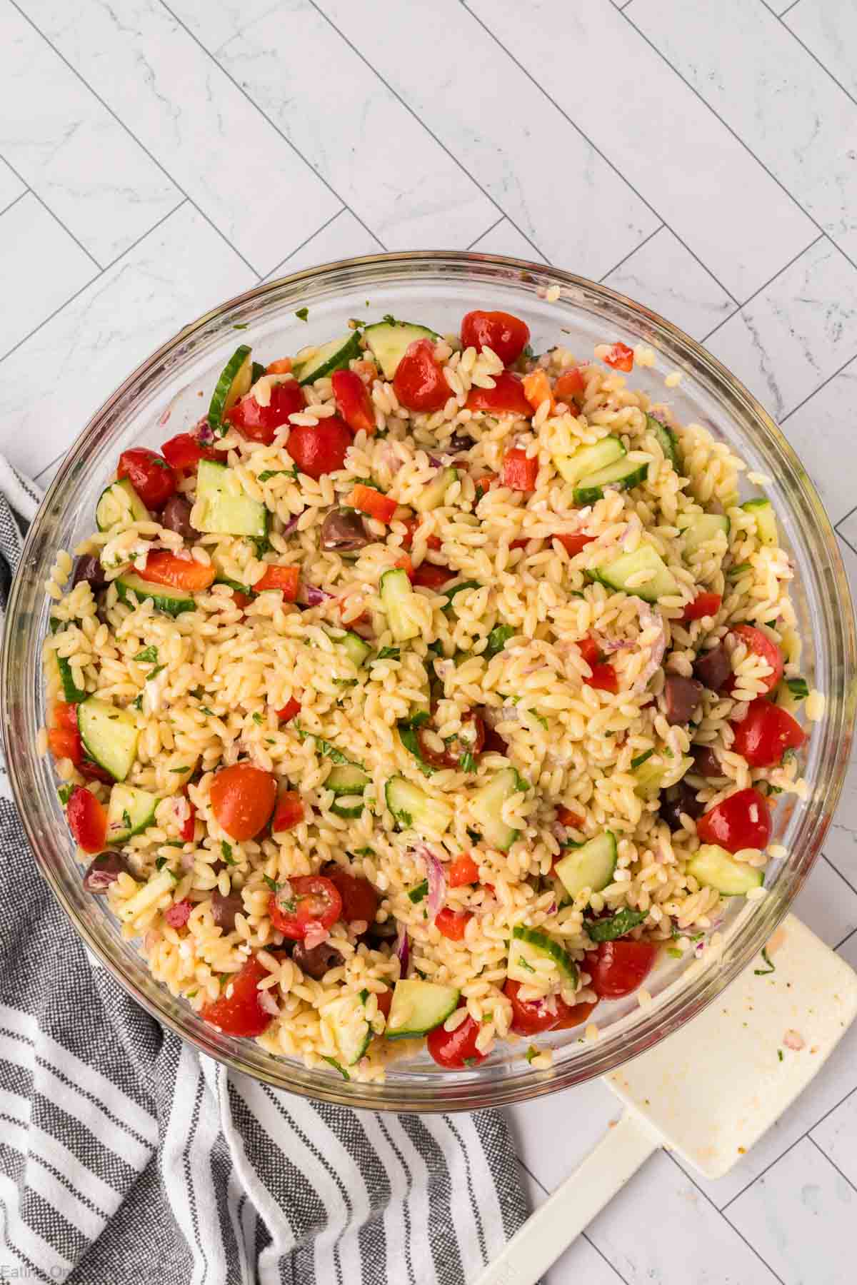 A glass bowl filled with an orzo pasta salad featuring diced cucumbers, halved cherry tomatoes, red onions, and black olives. The pasta salad is mixed with a light dressing and garnished with chopped herbs. A striped cloth and white spatula are positioned beside the bowl.