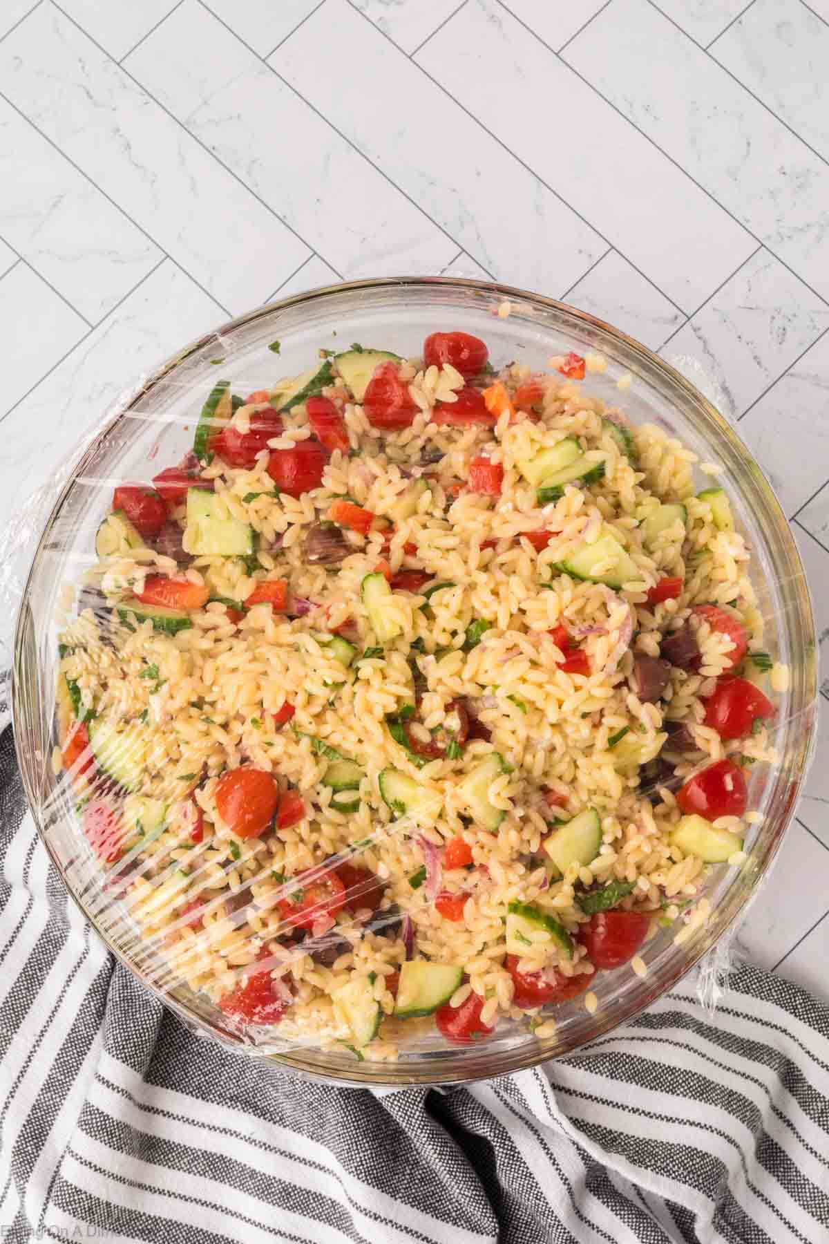 A glass bowl filled with orzo pasta salad, including ingredients like cherry tomatoes, cucumber slices, and finely chopped herbs, covered with plastic wrap. The bowl rests on a black and white striped cloth on a tiled surface.