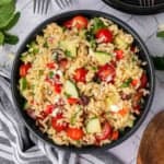 A bowl of orzo pasta salad with cherry tomatoes, cucumber slices, black olives, red onions, and crumbled feta cheese. The pasta salad is garnished with fresh parsley and placed on a white marble surface next to a striped cloth and utensils.